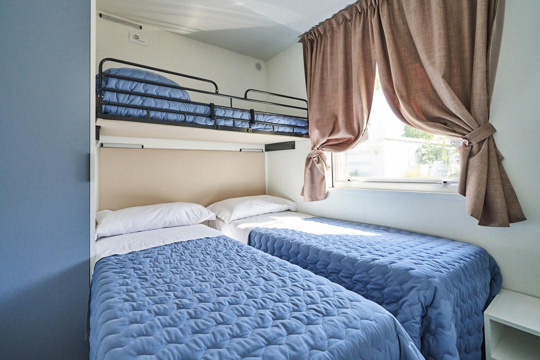 Room with bunk and single beds, blue bedspreads, window with brown curtains.